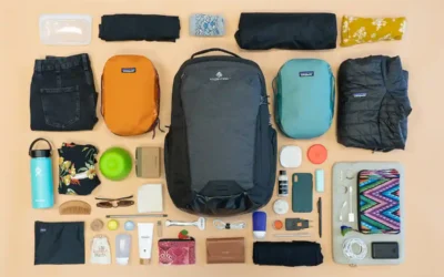 A Complete Guide of Best Travel Essentials for Anxiety A neatly arranged travel packing layout includes a black backpack, two smaller packing cubes (one orange, one teal), a rolled jacket, various clothing items, toiletries, a water bottle, a wallet, sunglasses, a phone, and other travel essentials on a beige background. Travel Essentials for Anxiety