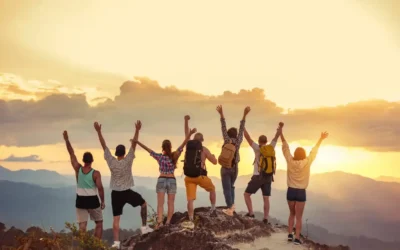 The Ultimate Guide to Solo Travel Groups for Independent Explorers Six people stand on a rocky hilltop at sunset, facing away from the camera and holding their arms up in celebration. They are dressed in casual outdoor clothing, some with backpacks, surrounded by a scenic landscape of distant mountains and a dramatic sky. Solo Travel Groups