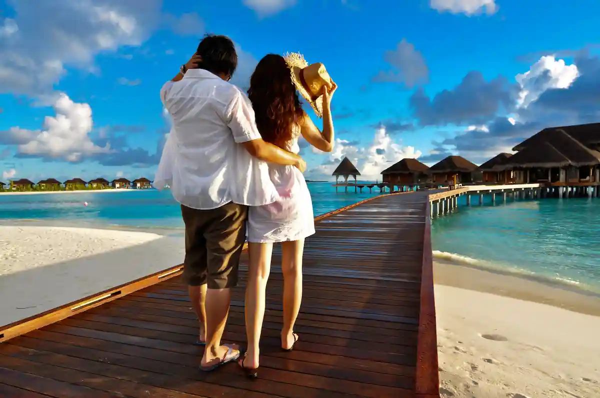 15 Romantic Things To Do in New Haven For Couples in 2024 2 A couple dressed in summer clothing walks arm-in-arm on a wooden boardwalk above turquoise waters. They are heading towards thatched-roof huts against a backdrop of a blue sky with scattered clouds. The woman holds a straw hat in her hand.