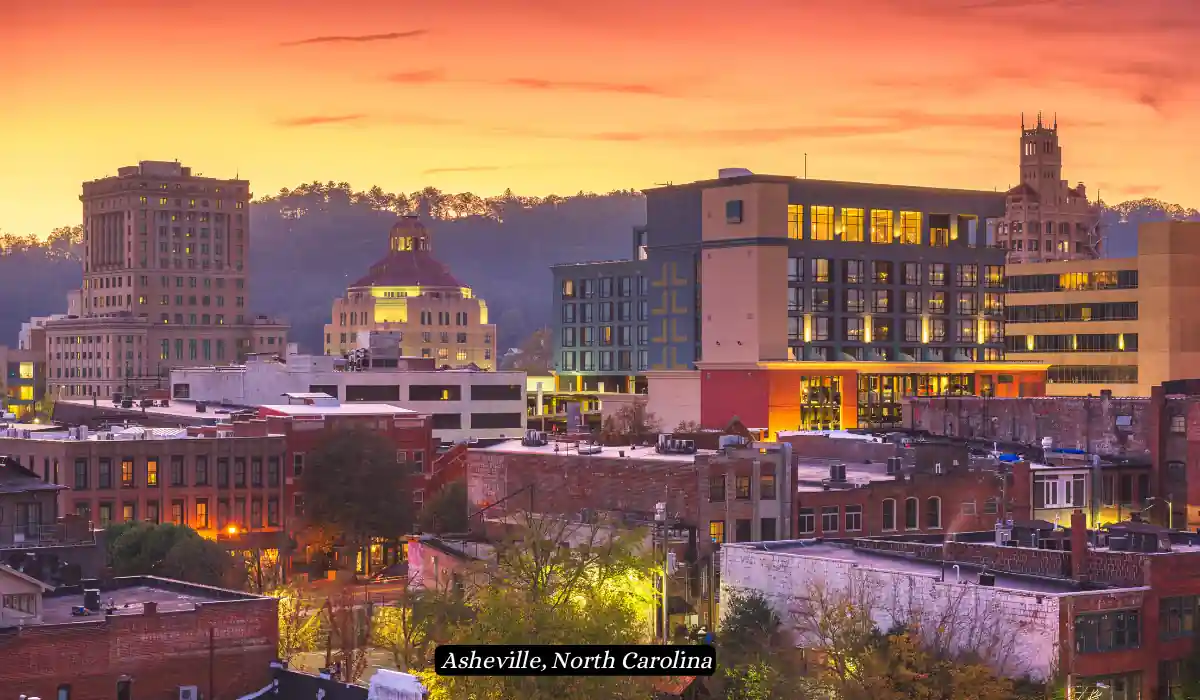 12 Best Places to Visit in November for Couples 2025 12 A scenic view of Asheville, North Carolina at sunset, showcasing a mix of historic and modern buildings under a vibrant orange sky. Trees and rooftops are visible in the foreground, with a label indicating the location at the bottom center of the image.