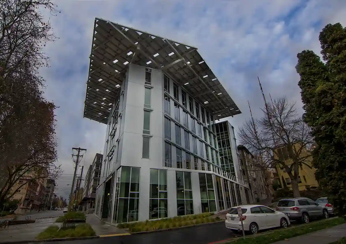 15 Romantic Things To Do in New Haven For Couples in 2024 7 A modern, multi-story building with large windows and a distinctive overhanging rooftop covered with solar panels. The structure is situated on a corner in an urban area with cars parked nearby and trees lining the street, under a cloudy sky.