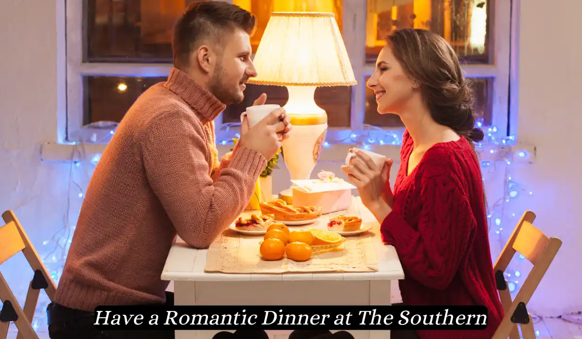 Top 15 Romantic Things to do in Nashville Tennessee for Couples 6 A couple sits across from each other at a small, white table, enjoying a cozy dinner. The table is adorned with an assortment of food and lit by a warm lamp, creating an intimate atmosphere. The image caption reads, "Have a Romantic Dinner at The Southern.