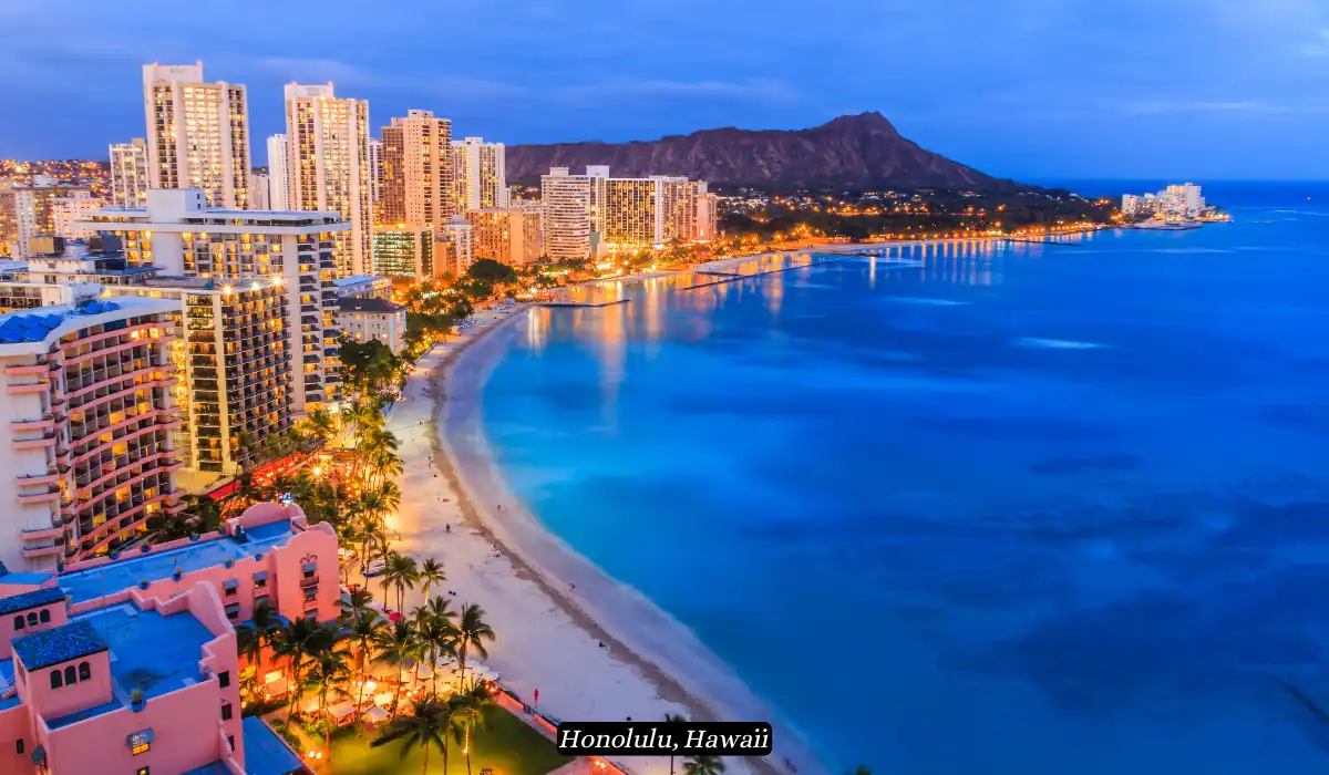 12 Best Places to Visit in November for Couples 2025 7 Aerial view of Waikiki Beach in Honolulu, Hawaii, at dusk. The city skyline is lit up with numerous buildings, while the calm waters of the Pacific Ocean reflect the lights. The iconic Diamond Head crater is visible in the background.
