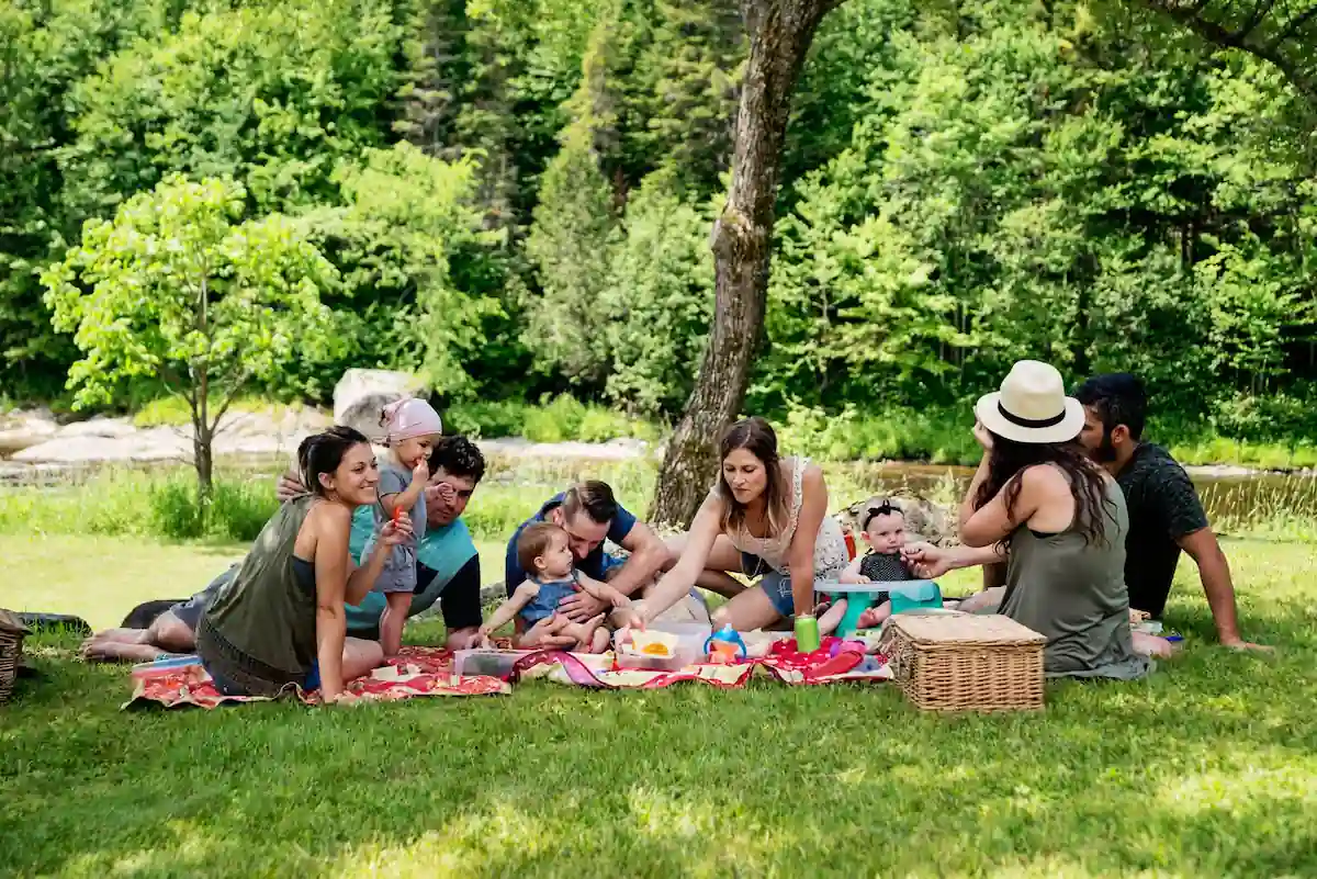 15 Romantic Things To Do in New Haven For Couples in 2024 4 A group of people, including adults and children, are sitting on a blanket having a picnic in a lush, green park. They are surrounded by trees and a pond is visible in the background. There is food spread out on the blanket and a wicker basket nearby.