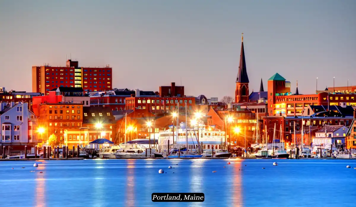 12 Best Places to Visit in November for Couples 2025 8 A vibrant evening view of Portland, Maine, showcasing the illuminated cityscape with a mix of modern and historic buildings, docked boats in the harbor, and the calm blue waters in the foreground. A prominent church steeple adds to the picturesque scene.