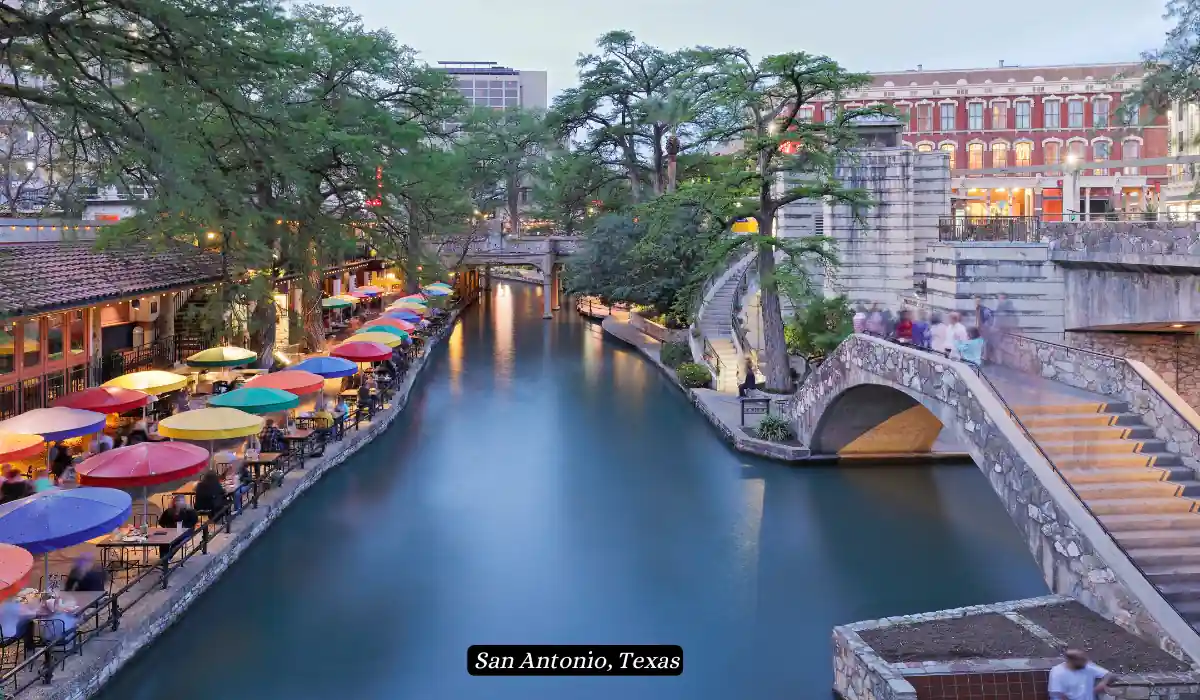 12 Best Places to Visit in November for Couples 2025 11 A serene evening view of the San Antonio River Walk in Texas. The scene includes a calm river, colorful umbrellas shading outdoor tables on the left side, lush trees, and quaint stone bridges. The background features historic buildings and softly lit pathways.