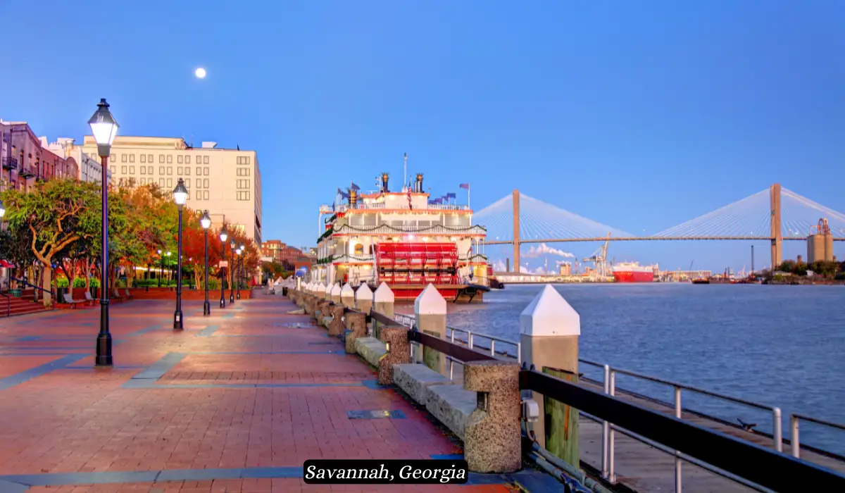 12 Best Places to Visit in November for Couples 2025 16 A scenic view of Savannah, Georgia's riverfront during twilight, featuring a historic paddleboat docked by the water, a brick-paved walkway with street lamps and benches, and the Talmadge Memorial Bridge in the background. The moon is visible in the clear sky.