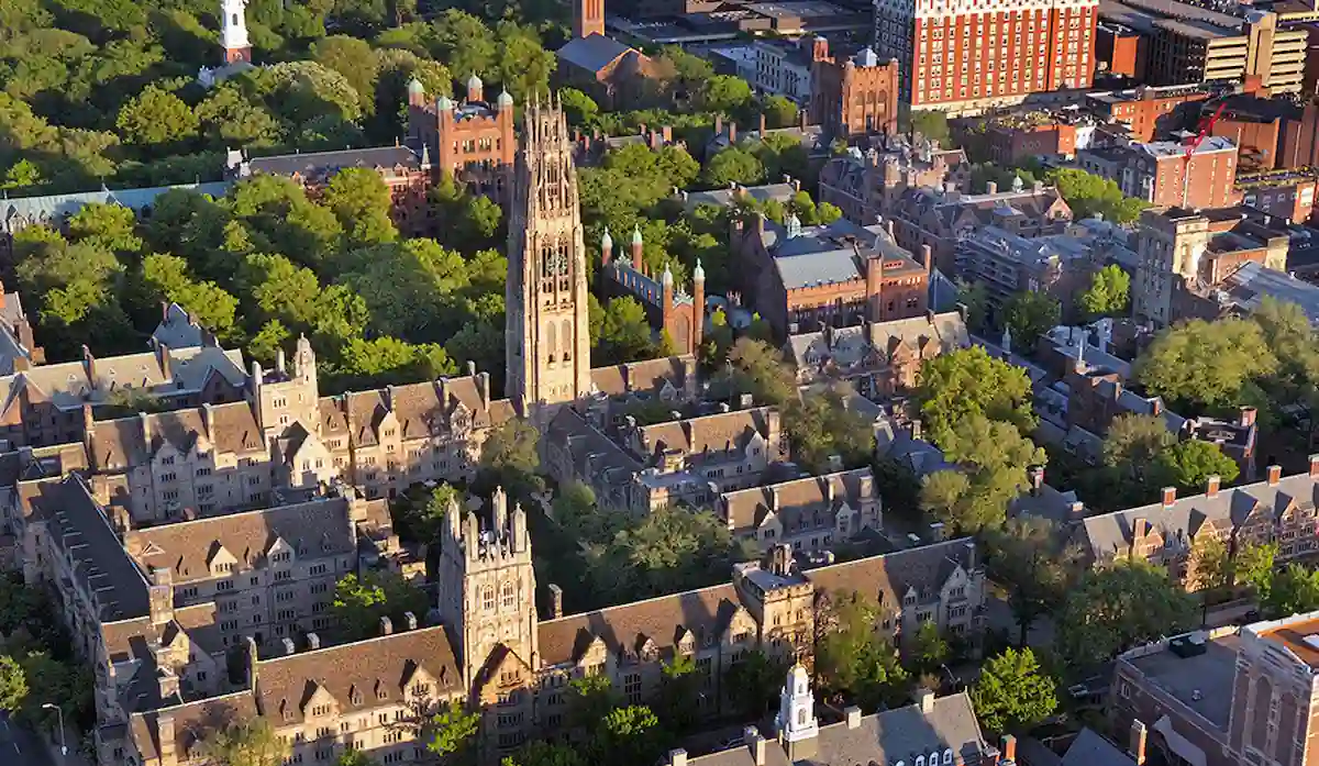 15 Romantic Things To Do in New Haven For Couples in 2024 3 Aerial view of a historic university campus with numerous Gothic-style buildings, prominent spires, and lush green trees. A tall bell tower stands at the center, surrounded by various academic buildings and residential areas.