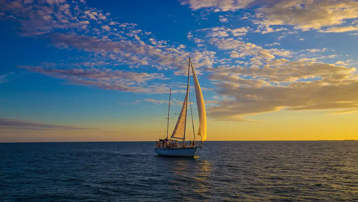 15 Romantic Things To Do in New Haven For Couples in 2024 5 A sailboat with white sails glides on a calm sea during sunset. The sky is vibrant with hues of blue, yellow, and orange, and scattered with fluffy clouds. The boat's silhouette is prominently visible against the colorful sky.