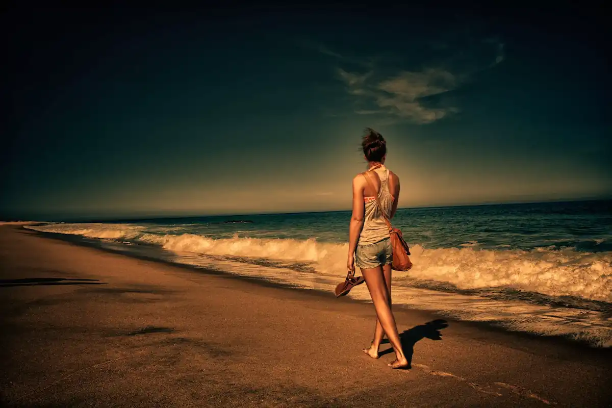 15 Romantic Things To Do in New Haven For Couples in 2024 14 A person wearing shorts and a tank top walks barefoot on a sandy beach at sunset. They hold a pair of sandals in one hand and have a bag slung over their shoulder. The ocean waves roll onto the shore, and the sky has deep hues of blue and orange.