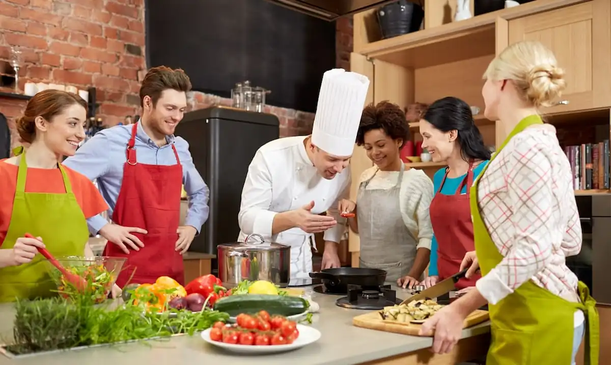 15 Romantic Things To Do in New Haven For Couples in 2024 15 A chef in a white uniform and hat is demonstrating cooking techniques to a group of five adults in a kitchen. They are all wearing colorful aprons and look engaged. Various fresh vegetables are on the counter, and a woman is chopping vegetables while others watch.