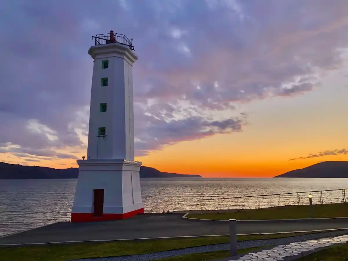 15 Romantic Things To Do in New Haven For Couples in 2024 9 A solitary white lighthouse with green windows stands on the edge of a paved road overlooking a calm body of water during sunset. The sky is a blend of orange, purple, and blue hues, and dark silhouettes of distant landmasses are visible in the background.