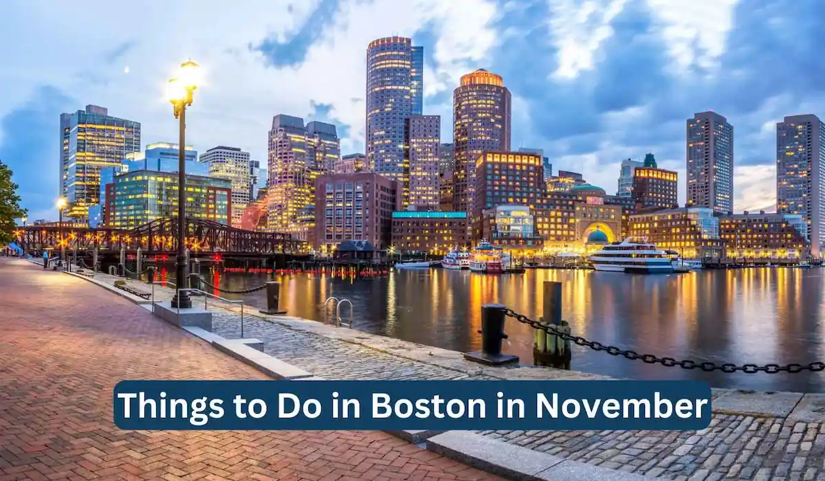 A scenic view of Boston's waterfront in the evening, with illuminated skyscrapers reflecting on the water. A brick-paved path runs along the waterfront with streetlights casting a warm glow. Text overlay reads, "Things to Do in Boston in November.