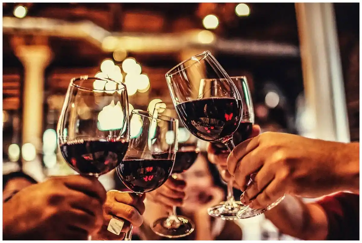 15 Romantic Things To Do in New Haven For Couples in 2024 12 A group of people toasting with red wine glasses in a warmly lit, cozy setting. The background is blurred, featuring soft lights and what appears to be a rustic interior, enhancing the intimate and celebratory atmosphere.