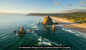A scenic view of Cannon Beach, Oregon, featuring the iconic Haystack Rock surrounded by ocean waves. The coastline stretches with sandy beaches and lush green hills in the background under a blue sky. "15+ Romantic Things to Do in Cannon Beach for Couples"