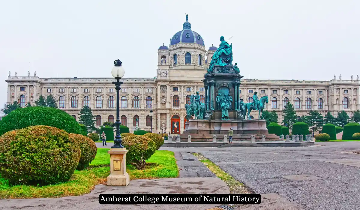 15 Free Romantic Things to Do in Massachusetts for Couples 8 A large, ornate building with a dome stands in the background, with a detailed statue surrounded by greenery in front. A lamp post and shrubs line the walkway. The image label reads "Amherst College Museum of Natural History.