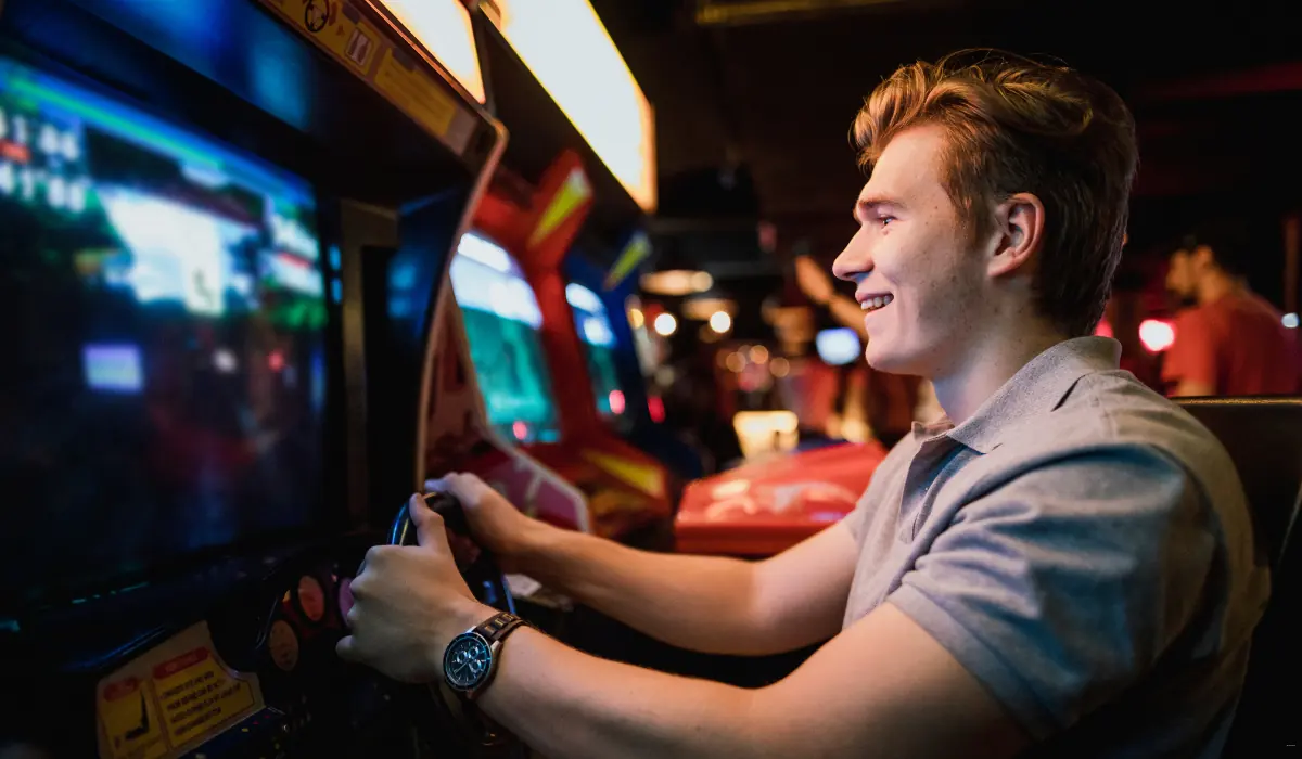 40 amazing Cute Date Ideas in Columbus, Ohio for Couples in 2025 31 A young man with short light brown hair is smiling while playing an arcade racing game. He is holding a steering wheel, and the arcade screens are brightly lit. The background is dimly lit with some people and more arcade machines visible.