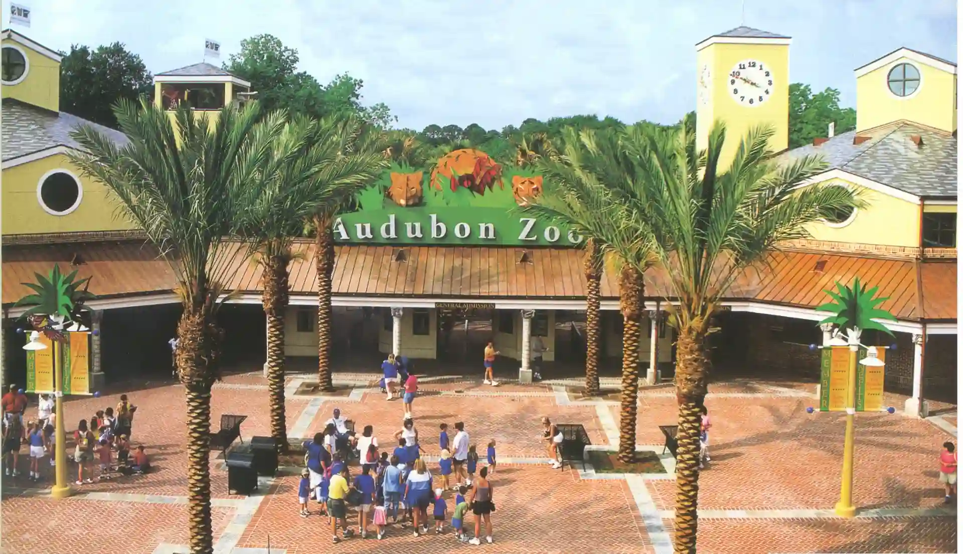 The image shows the entrance to Audubon Zoo, featuring a clock tower, palm trees, and a colorful, busy plaza with visitors. The zoos name is prominently displayed on a yellow and green sign above the entrance.
