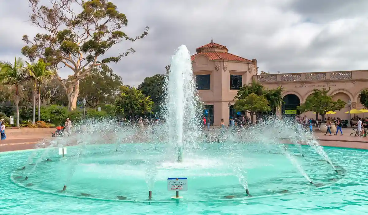 12+ Best Things to do in San Diego for Couples 2025 5 A large fountain with vibrant water jets in a circular basin is set in a park. Trees and a historic building with arches are in the background. People are gathered around under a cloudy sky.