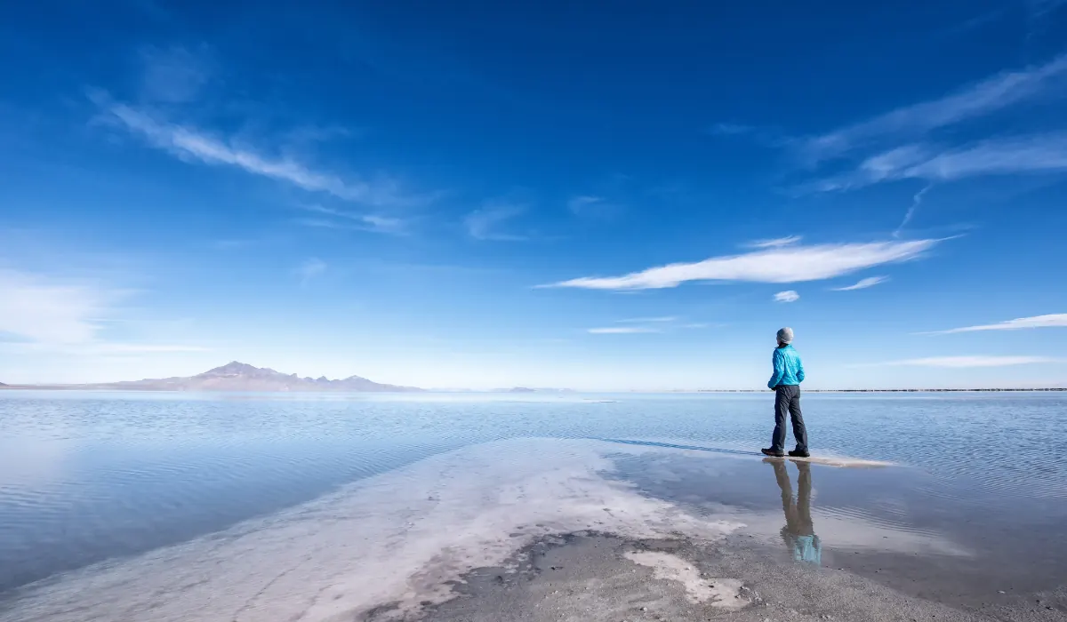 Top 15 Fun $ Unique Places That Don't Feel Real in the USA 2025 2 A person in a blue jacket and black pants stands on a narrow strip of land, gazing across a vast, reflective salt flat under a bright blue sky with scattered clouds. Distant mountains are visible on the horizon.