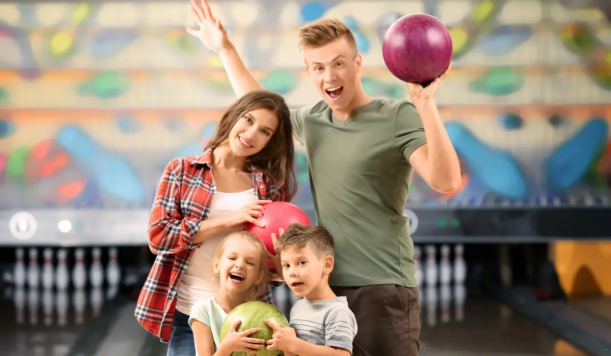 40 amazing Cute Date Ideas in Columbus, Ohio for Couples in 2025 18 A smiling family of four at a bowling alley, each holding a bowling ball. The father and mother are standing behind their two young children. The background shows a row of bowling pins and a colorful mural.