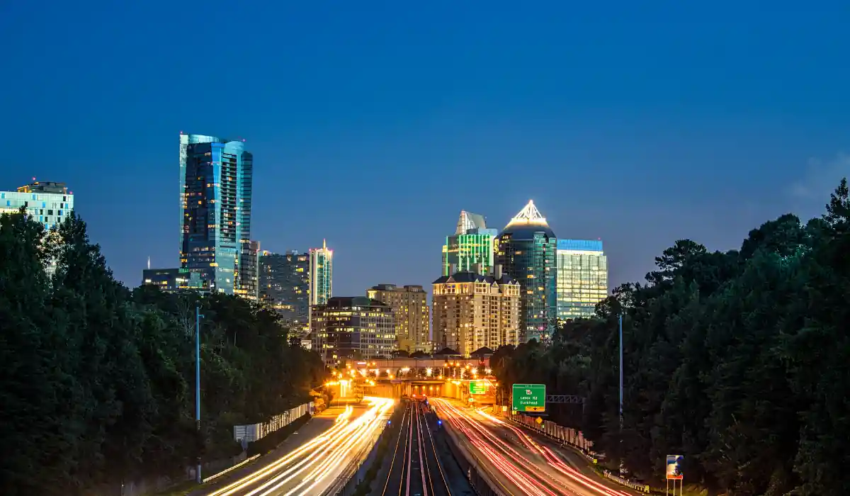 Top 15+ Free Things to do in Atlanta for Couples 2025 14 A nighttime cityscape featuring a highway with light trails from moving vehicles. Tall buildings with illuminated windows rise against a deep blue sky. Trees line the sides of the highway, framing the urban skyline.