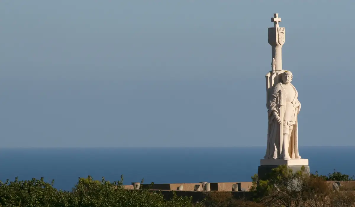 12+ Best Things to do in San Diego for Couples 2025 7 A large statue of a figure holding a cross stands against a clear blue sky. The statue overlooks a distant body of water, with greenery in the foreground.
