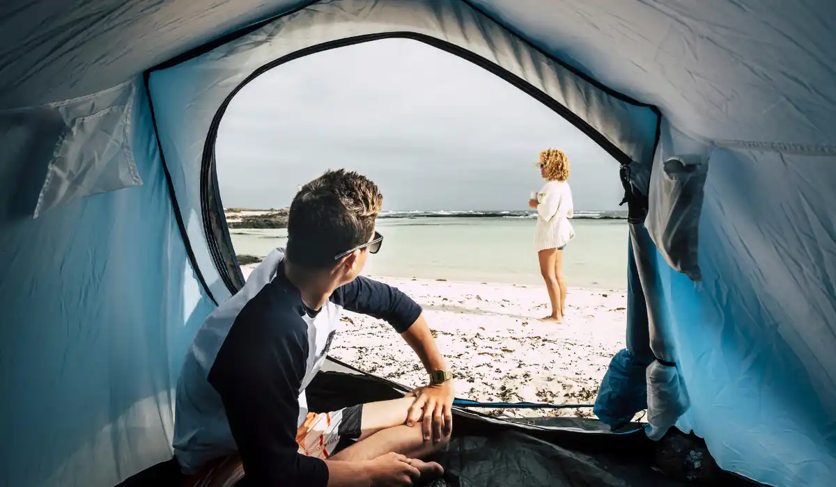 20 Most Romantic Things to do in California for Couples 20 View from inside a tent looking out at a beach scene. A person sits inside the tent, gazing at another person standing on the sandy shoreline. The ocean and overcast sky are visible in the background.