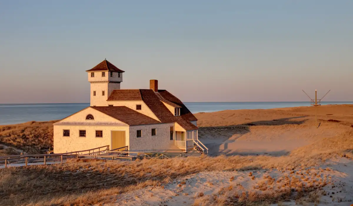 A rustic, white coastal house with a small tower stands amidst sandy dunes and tall grasses during sunset. The ocean is visible in the background under a clear sky.
