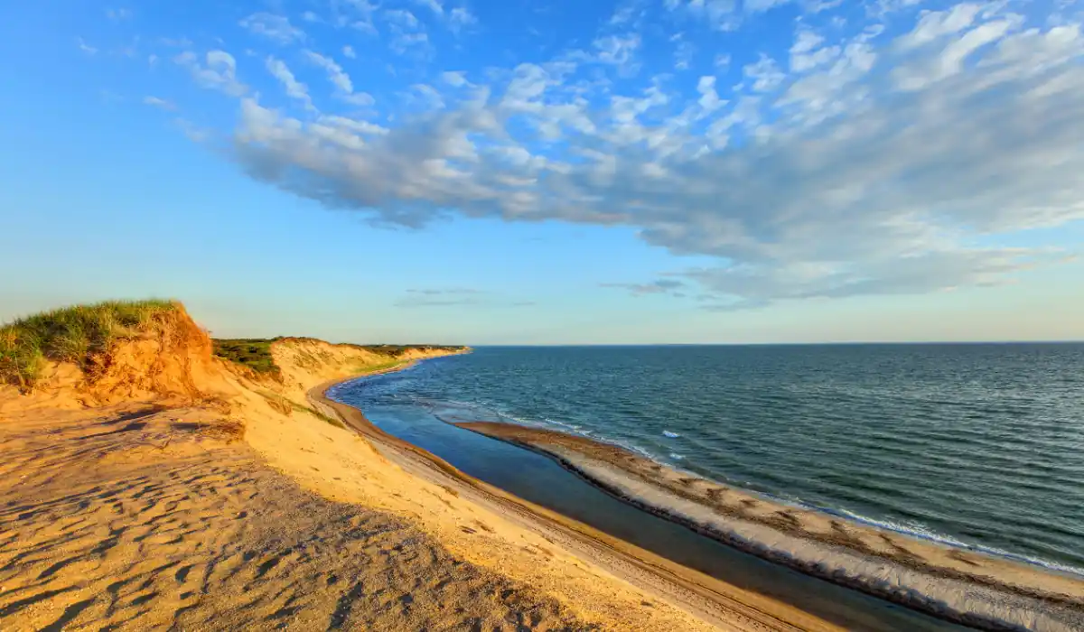 A scenic coastal view with golden sand dunes on the left, a narrow strip of land dividing a small river and the ocean. The sky is mostly clear with a few scattered clouds, and the sunlight casts warm tones over the landscape.
