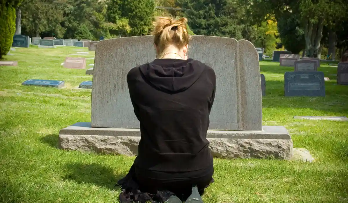 The 15 Best Things to Do in New Orleans for Couples 15 A person kneels in front of a gravestone in a cemetery, wearing black clothing. The sun is shining, and there are several other gravestones in the background amidst greenery and trees.