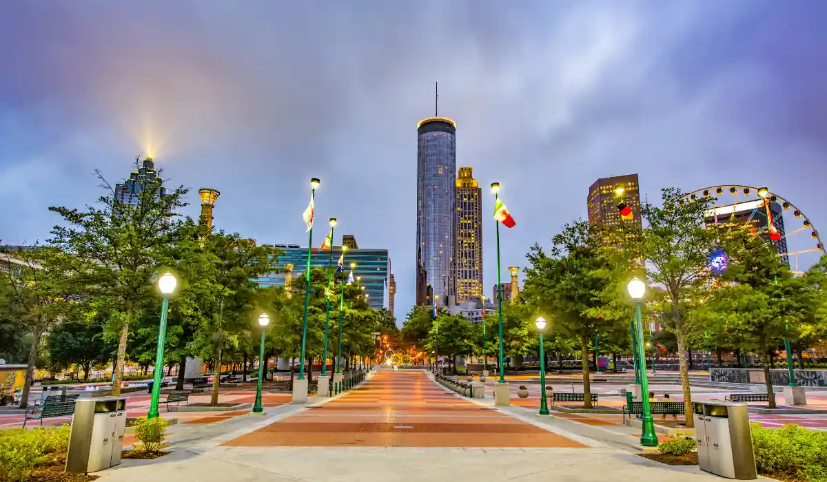 Top 15+ Free Things to do in Atlanta for Couples 2025 3 A vibrant cityscape featuring a wide, lit pathway lined with trees and lampposts leading towards tall skyscrapers. The Ferris wheel on the right adds a touch of whimsy to the urban setting under a cloudy sky.