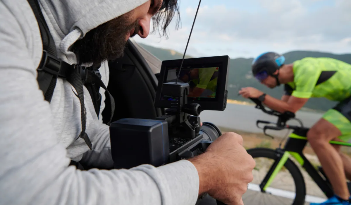 40 amazing Cute Date Ideas in Columbus, Ohio for Couples in 2025 37 A person in a hooded sweatshirt films a cyclist in a neon green outfit riding a bike. The cyclist is racing on a road beside a body of water, with mountains in the background. The camera's screen displays the cyclist's image.