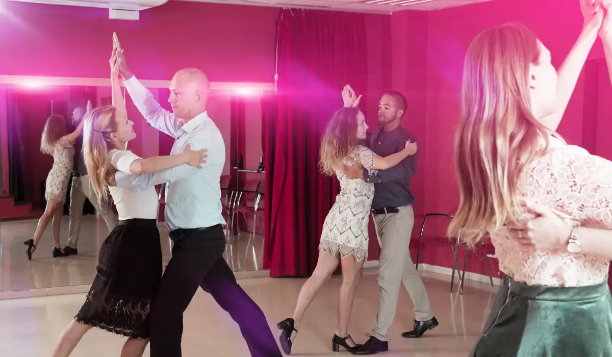 40 amazing Cute Date Ideas in Columbus, Ohio for Couples in 2025 28 Four couples are ballroom dancing in a studio with red walls and mirrors. They are gracefully moving in pairs, with a pink light illuminating the room, and chairs visible in the background.