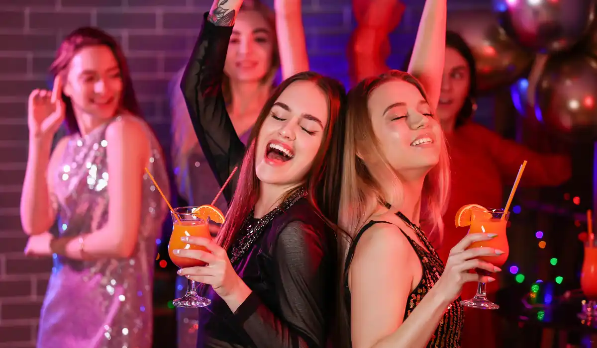 40 amazing Cute Date Ideas in Columbus, Ohio for Couples in 2025 21 Two women with long hair are smiling and dancing back-to-back, holding orange drinks with straws. They're surrounded by others at a lively party, with colorful lights and balloons in the background.