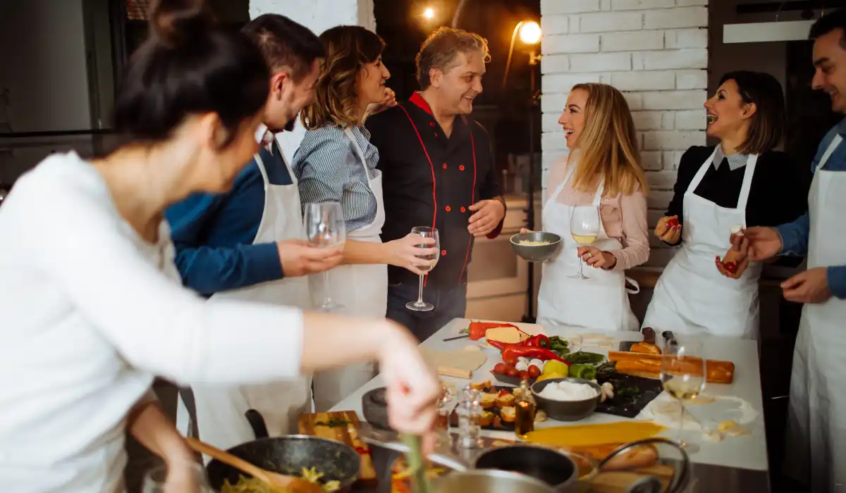 40 amazing Cute Date Ideas in Columbus, Ohio for Couples in 2025 20 A group of people in a kitchen setting, wearing aprons, are gathered around a table filled with various ingredients and dishes. They are smiling and conversing, with one person holding a glass of wine. Cooking utensils and food items are visible on the table.