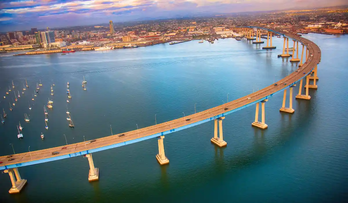 12+ Best Things to do in San Diego for Couples 2025 11 Aerial view of a long, curved bridge spanning over a body of water. Sailboats are anchored in the calm water. The coastline cityscape is visible in the background under a partly cloudy sky.