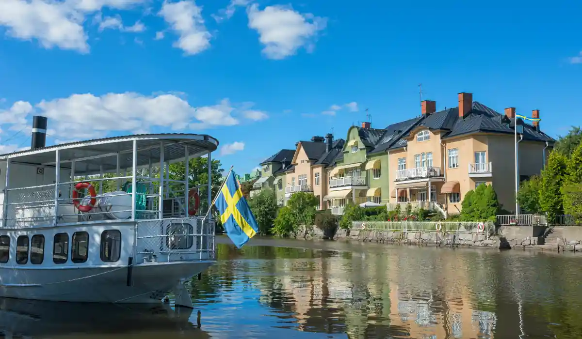 The 15 Best Things to Do in New Orleans for Couples 14 A scenic view of a river with a boat displaying a Swedish flag. In the background, colorful houses line the riverbank under a clear blue sky with scattered clouds.