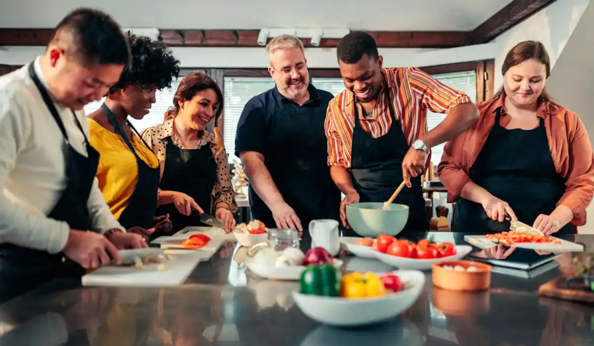 The 15 Best Things to Do in New Orleans for Couples 16 A diverse group of six people in aprons are in a kitchen, smiling as they prepare food together. Some chop vegetables on cutting boards while others mix ingredients in a bowl. Various fresh vegetables are visible on the counter.