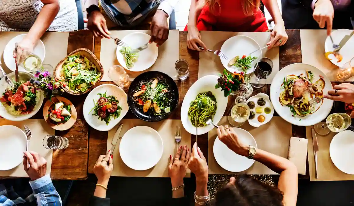40 amazing Cute Date Ideas in Columbus, Ohio for Couples in 2025 25 A group of people is seated around a wooden table filled with various dishes, including salads, pasta, and roasted meat. Hands are reaching for food, and there are drinks on the table, suggesting a communal meal or gathering.