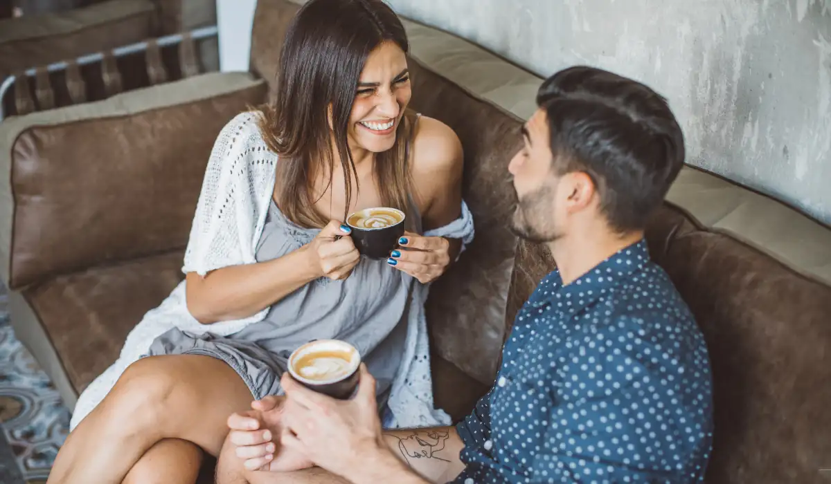 40 amazing Cute Date Ideas in Columbus, Ohio for Couples in 2025 5 A woman and a man sit on a brown couch, smiling at each other while holding cups of cappuccino. The woman wears a gray dress and a white shawl, and the man wears a blue polka dot shirt. They appear to be enjoying a casual conversation.