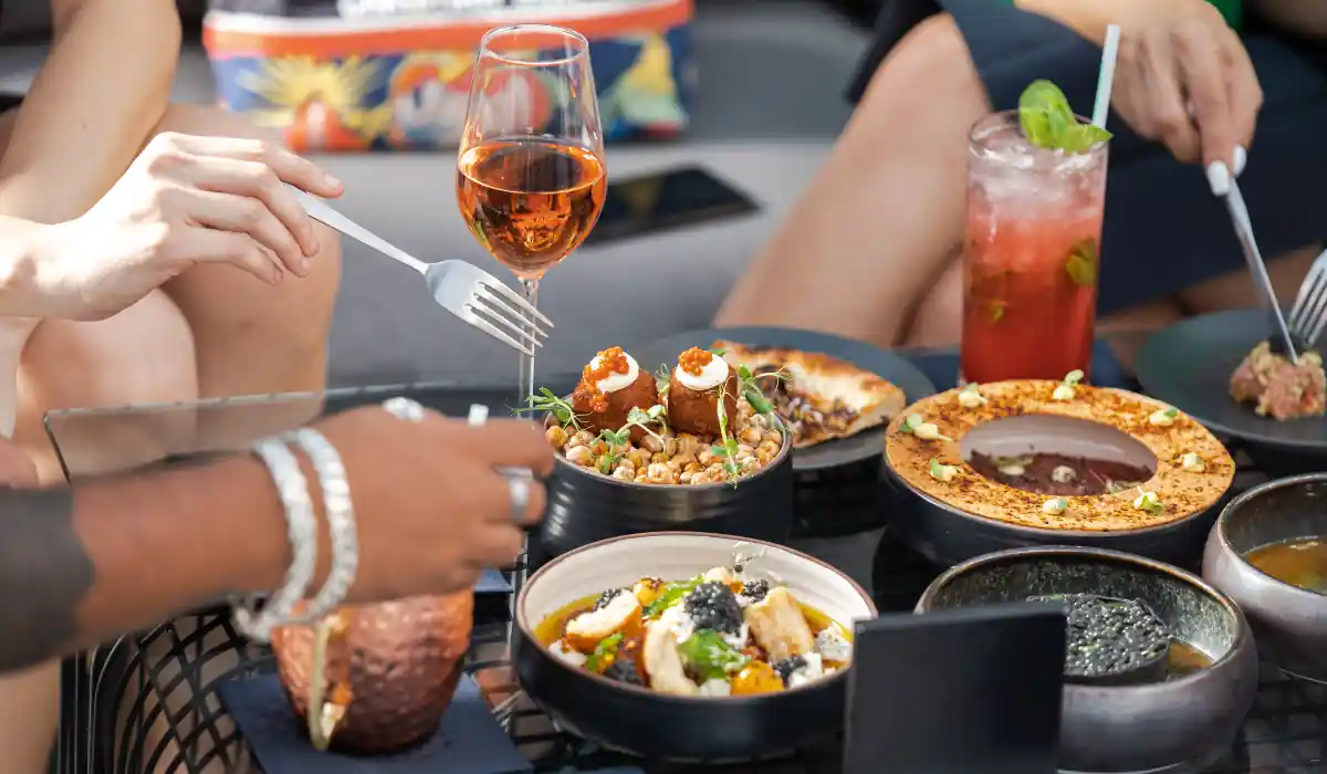 40 amazing Cute Date Ideas in Columbus, Ohio for Couples in 2025 29 A table setting featuring a variety of dishes, including a bowl of chickpeas, a plate of appetizers, and several glasses of drinks. Three people are enjoying the meal, reaching for food with forks. A glass of rosé wine is prominently displayed.