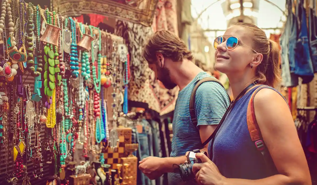 The 15 Best Things to Do in New Orleans for Couples 19 A woman wearing sunglasses and holding a camera stands next to a man in a striped shirt, both browsing vibrant and colorful jewelry at a market stall. The scene captures a lively atmosphere with diverse accessories on display.