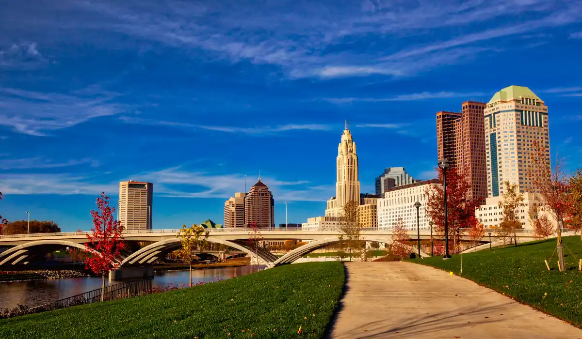 40 amazing Cute Date Ideas in Columbus, Ohio for Couples in 2025 3 A vibrant cityscape of downtown Columbus, Ohio, featuring modern skyscrapers against a clear blue sky. A paved pathway runs alongside lush green grass and colorful autumn trees near a river, with an arched bridge in the background.