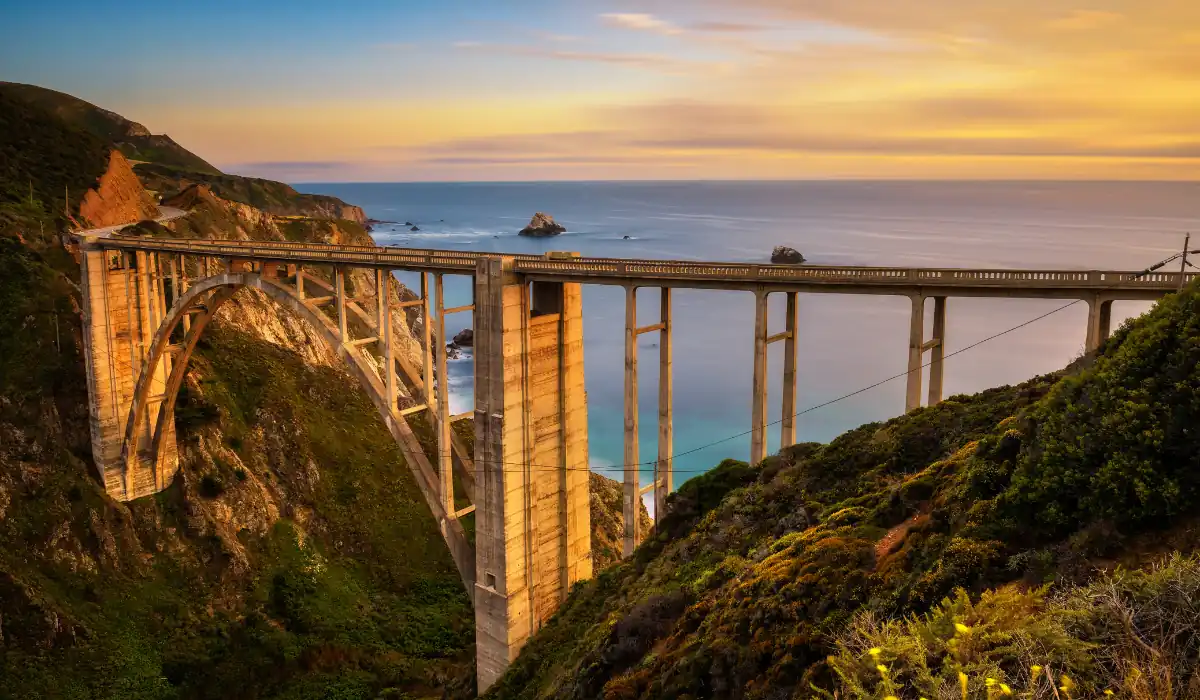 20 Most Romantic Things to do in California for Couples 11 A scenic view of Bixby Creek Bridge during sunset. The bridge spans across a deep canyon with rugged cliffs and lush greenery. The golden sky reflects on the calm ocean waters in the background, creating a tranquil atmosphere.