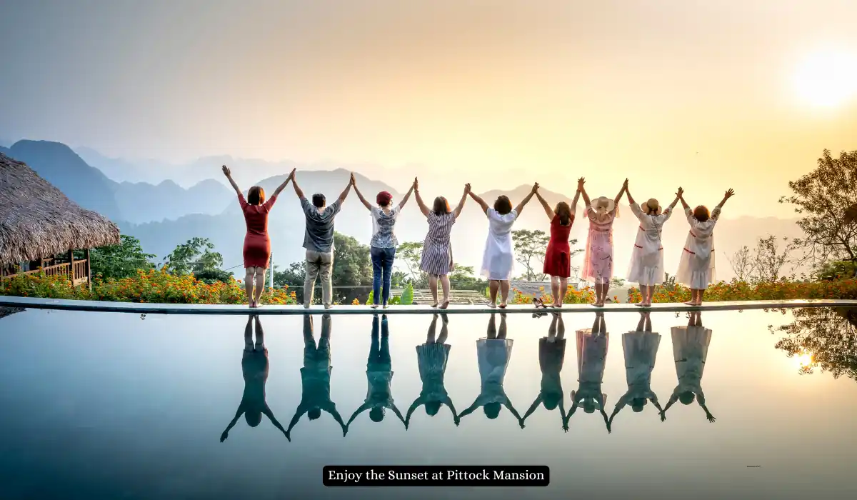 Top 15+ Romantic Things to do in Portland for Couples 9 A group of people stand hand in hand, facing a scenic sunset. Their reflections are visible in a calm body of water in front of them. Lush greenery and distant mountains frame the horizon. A caption reads, "Enjoy the Sunset at Pittock Mansion.