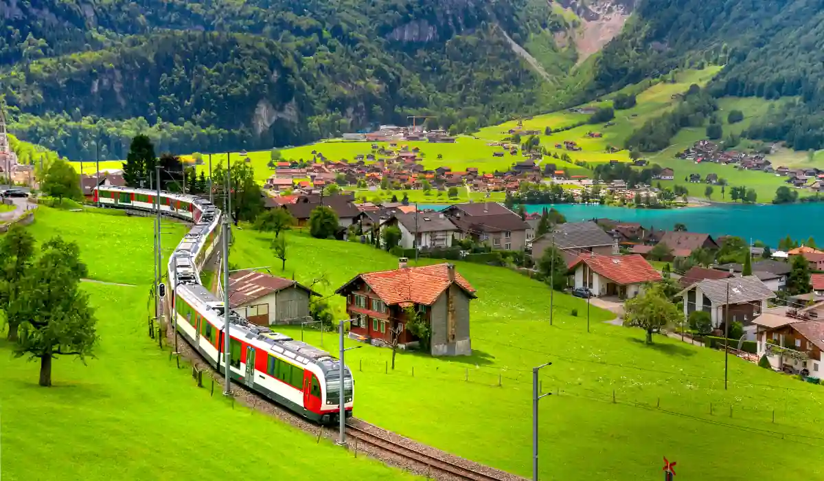 40 amazing Cute Date Ideas in Columbus, Ohio for Couples in 2025 32 A passenger train travels through a lush green valley with scattered houses. In the background, a turquoise lake rests at the base of forested mountains. The sky is partly cloudy, adding depth to the picturesque landscape.