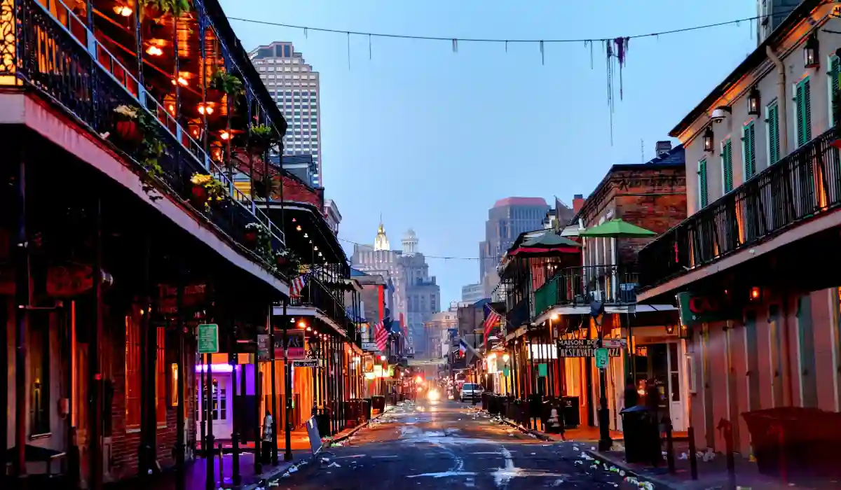 The 15 Best Things to Do in New Orleans for Couples 7 A vibrant street scene in New Orleans featuring illuminated buildings with balconies and streetlights under a dusky sky. The street is wet, reflecting the colorful lights, and skyscrapers are visible in the background.