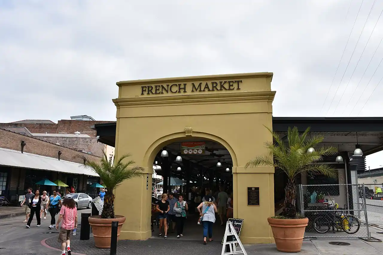 The image shows the entrance to the French Market, featuring an archway with French Market written on it. People are walking in and out of the market area, and there are palm trees and bicycles nearby. The sky is cloudy.
