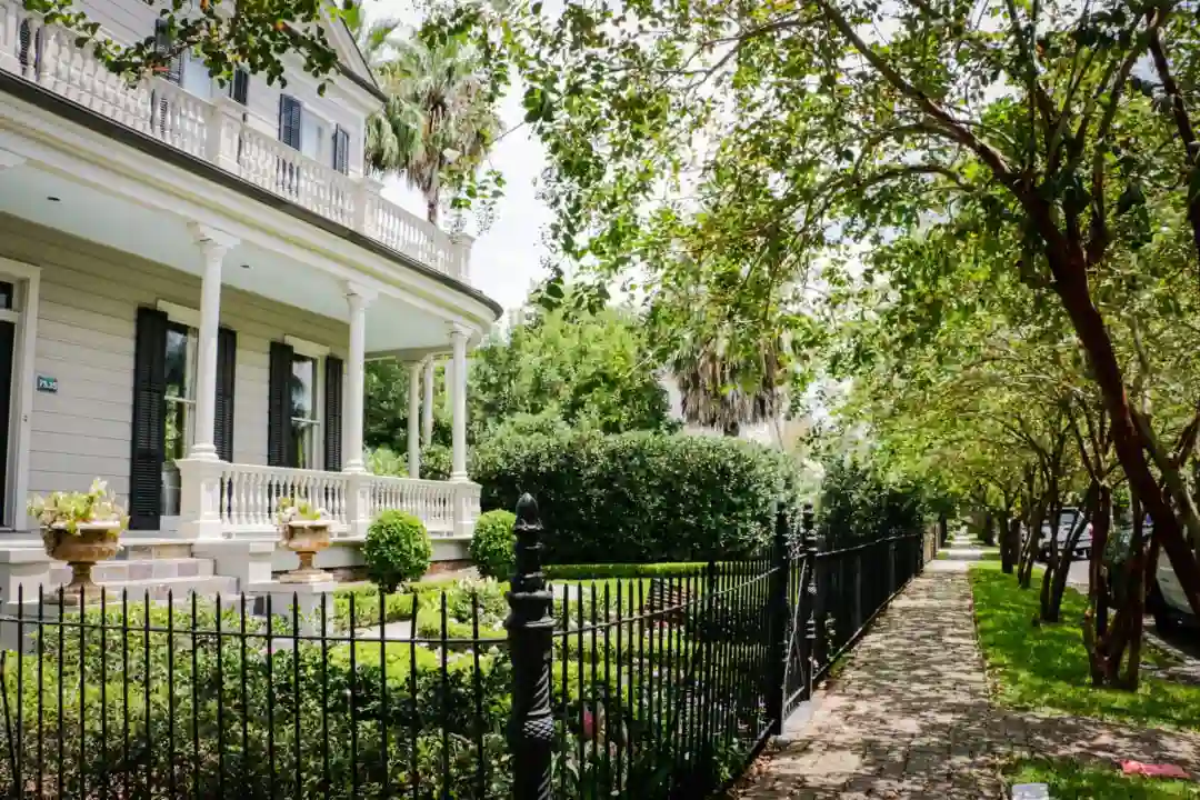 A grand, two-story white house with a wraparound porch is surrounded by lush greenery and a manicured lawn. A black iron fence lines the sidewalk, which is shaded by trees under a clear sky.
